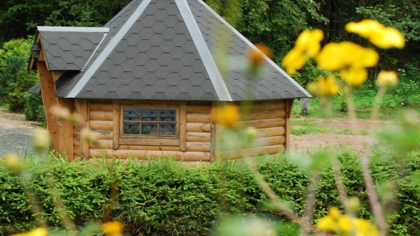 Kleine runde Holzhütte mit schwarzem Dach, Fenster und Eingang hinter Blumen und Hecke vor Wald