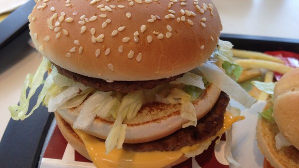 Großer Hamburger mit doppelt Fleisch, Salat und Käse auf Tablett. Auf dem Burger-Brötchen sind Sesam-Körner.