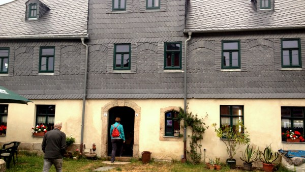 Hausferont mit 3 Etagen, denkmalgeschützt, viele Pflanzen. unten beige in der Mitte an der Wand und auf dem Dach Schiefer-Oberfläche. Grüne Fenster