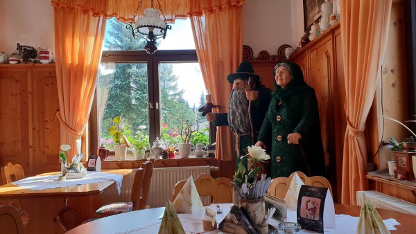 Urigeer Gastraum mit gedeckten Tischen. In der Ecke stehen Moosmann und Moosfrau. In der Mitte ein schön verziertes Fenster.