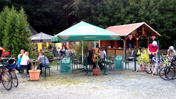 Platz vor dem Biergarten mit Fahrradständern an der Seite. In der Mitte Sonnenschirm und Angebotstafeln