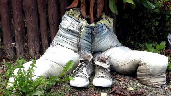 Hose als Blumentopf mit Schuhen als Deko-Elemente vor Holzzaun. Passend davor Unkraut.