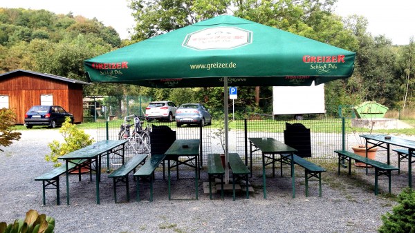 Sonnenschirm wirft Schatten auf Bierzeltgarnituren. Im Hintergrund stehen Autos auf einem Parkplatz