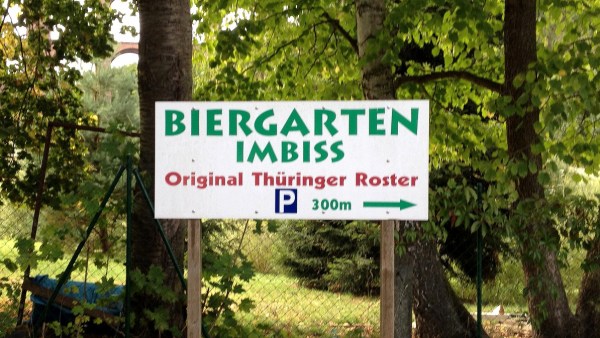 Weißes Schild vor Bäumen und Zaun mit Text: Biergarten Imbiss Original Thüringer Roster P 300 m