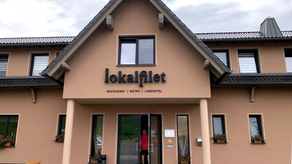 Helles terracottafarbenes Haus. 2 Etagen, Giebel, Eingang mit Schriftzug lokalfilet, darunter steht: Restaurant Bistro Landhotel. An der linken Seite eine Bank.