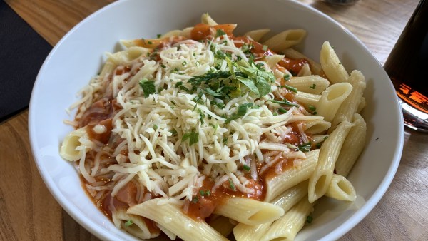 Teller auf Tisch, voll mit Nudeln, Tomatensoße und Käse, garniert mit Kräutern. Im Hintergrund sind eine Serviette und Trinkgläser zu sehen.