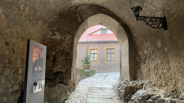 Rundbogen-Durchgang aus Stein. Links Infotafel, Rechts oben schwarze Lampe