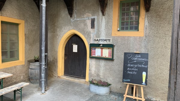 Orange umrandete Fenster und braune Spitzbogen-Tür aus Holz, Regenrinne, Schiefertafel mit Aufschrift Herzlich Willkommen Hausgebackener Kuchen Snacks, über einer Schautafel steht GASTSTÄTTE
