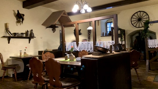 Innenraum einer Gaststube. Tisch, Stühle, Schrank aus dunklem Holz. Dekoration an der Wand: Auerhahn, Wagenrad.