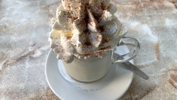 Glastasse mit weißer Schokolade auf Unterteller mit Löffel. Obendrauf eine Sahnehaube mit Schokoraspeln. Das Ganze auf einem marmorierten Tisch.