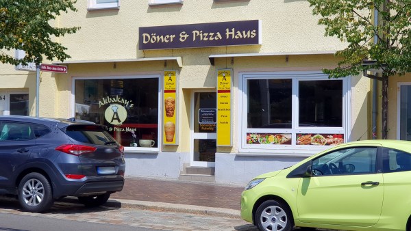 Döner-Imbiß in Haus mit zwei Schaufenstern und Eingang. Davor Fußweg und jeweils zur Hälfte zwei parkende Autos.