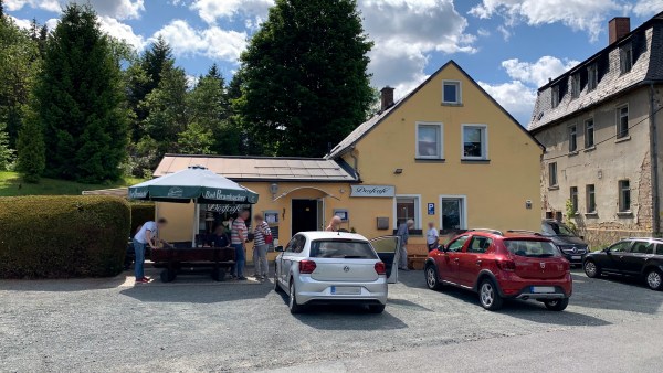 Silbernes und Rotes Auto parken vor gelbem Haus. Massive Holzbank und Bad Brambacher Sonnenschirm stehen vor dem Haus. Großer Platz davor. Rechts verfallenes Haus. Links Rasen, Hecke und Bäume. Schönes Wetter.