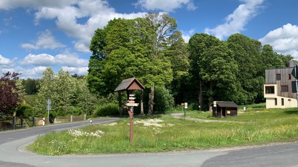 Straßenkurve um Wiese herum. In der Mitte wein Wegweiser. Hinten Bäume. Helles und sonniges Wetter.