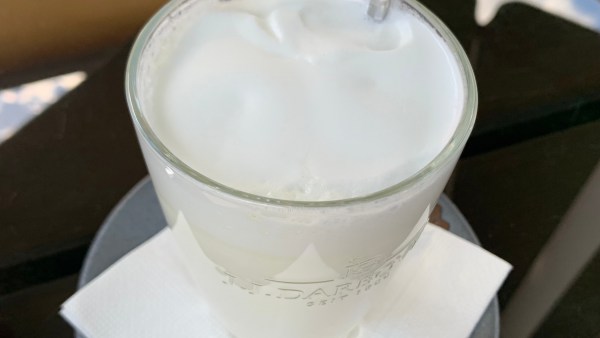 Weißes Heißgetränk in Glas mit Schaum auf Serviette auf Untertasse. Hinten sieht man noch ein wenig einen eingesteckten Löffel.