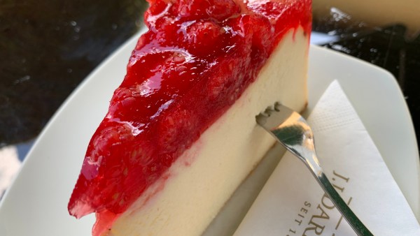 Quark-Sahne-Torte mit Himbeer-Topping auf weißem Teller neben Serviette. In dem Stück Kuchen steckt eine Kuchengabel.