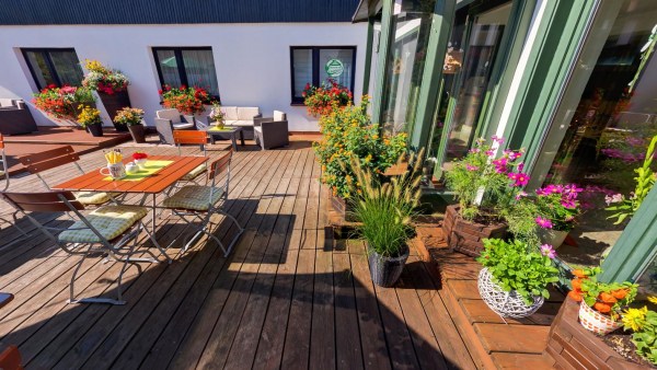 Holzterrasse mit Gartenmöbel, sitzecke, viele Blumentöpfe, hinten weiße Wand mit fenster, rechts Glasfassade