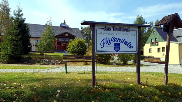 Willkommensschild auf Wiese vor Haus Flößerstube. Dazwischen Weg und Bank