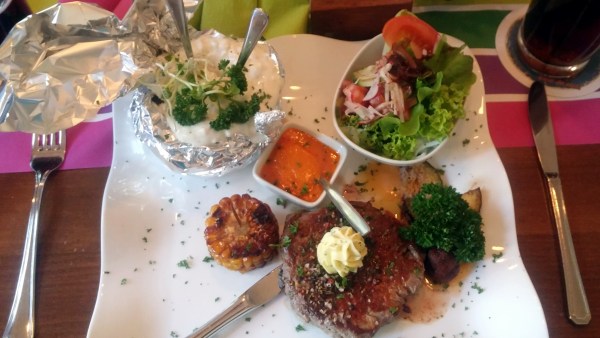 Rindersteak mit Salz und Butter auf welligem Teller, Dip, Salt und Ofenkartoffel mit Quark. Das ganze serviert auf einem Tisch zwischen Besteck.