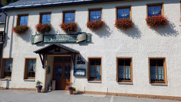 Hausfront von weißem Haus mit zwei Etagen. Oben alle Fenster mit Blumenstock und roten Blumen. Unten Fenster und Eingang zum Gasthaus.