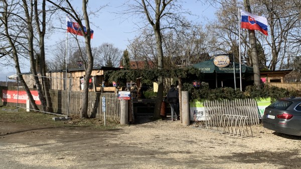 Vom Ascheplatz zum vom umzäunten Eingang. Im Gelände stehen Bäume, Holzhütten und ein Wernesgrüner-Bier-Schirm. Den den Seiten wehen jeweils slowakische Flaggen.
