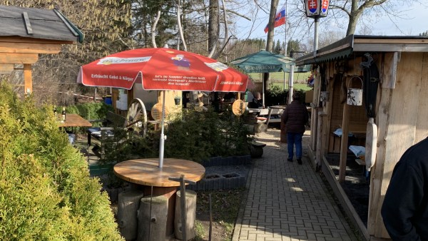 Gepflasterter Gang, zwischen offener Holzhütte. Im Hintergrund Büsche, Bäume, Gestaltungselemente mit slowakischen Symbolen. Eine Person läuft hinein. Sonnenschirme spenden Schatten.