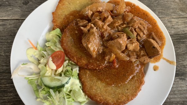 Weißer Teller auf Holztisch mit Bambes, Gulasch und Soße sowie etwas Rohkost, Salat, halbe Gurke, halbe Tomate und Dressing