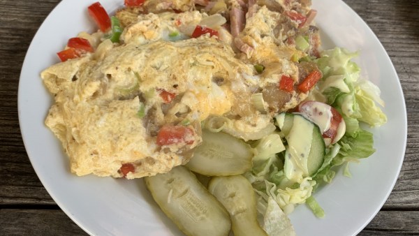 Weißer Teller auf Holztisch mit Ei-Omelette und Schinkenstreifen-Gemüse-Füllung sowie Gewürzgurken und Rohkost.