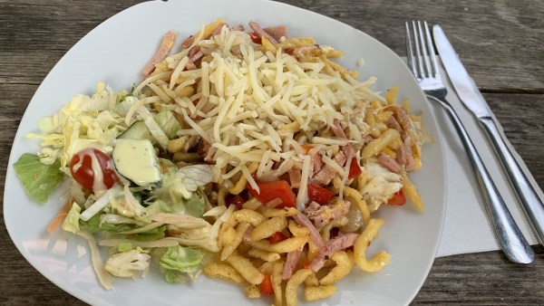 Weißer Teller auf Tisch mit Besteck und Serviette. Auf dem Teller Spätze, Paprika, Schinkenstreifen. Darauf geriebener Käse. Links Salat mit Tomate, Gurke und Dressing.