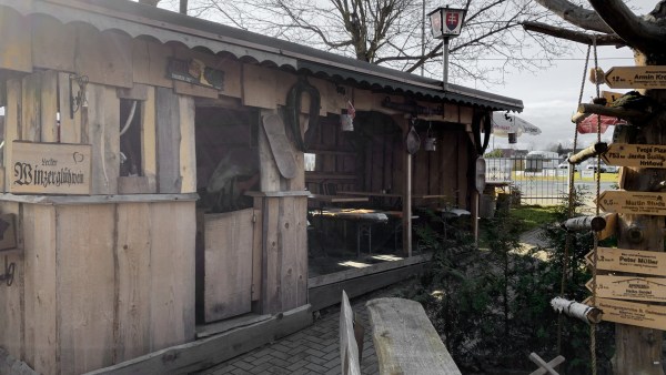 Holzhütte mit Weg und Bank davor. Rechts Hinweisschilder in Form von Wegweisern. Links Hinweis in altdeutscher Schrift auf Winzerglühwein.