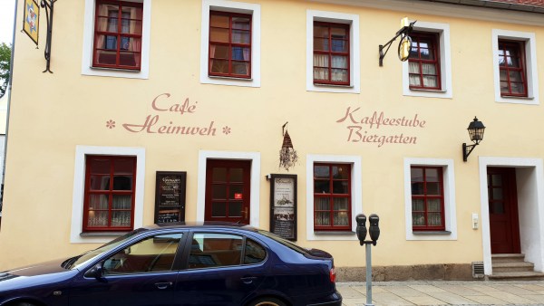 Zweistöckige Hausfront vom Cafe Heimweh. Im Vordergrund parkt ein blaues Auto