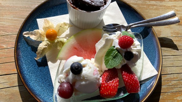 Auf einem Holztisch auf einem blauen Teller auf einer Serviette: Nachtisch mit Löffel zur rechten Seite ragend. Dessert bestehend aus einem Schoko-Küchlein in einem kleinen weißen Porzellan-Behälter, eine Kugel Joghurt-Eis mit Erdbeeren, Himbeere, einem Blatt Minze, einem Stück Melone und einer geöffneten Physalis