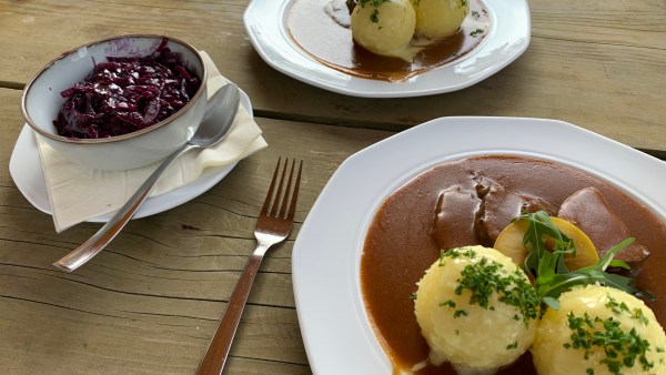 Auf einem Holz-Tisch zwei Teller mit Sauerbraten und Klößen, eine Gabel, eine Schüssel auf Serviette mit Rotkohl, daneben ein Löffel