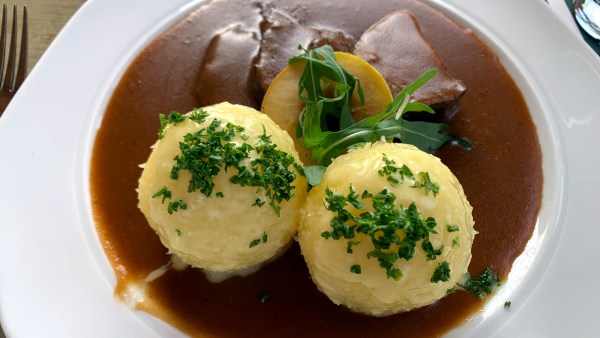 Teller mit Klößen, welche mit geriebener Petersilie garniert sind, neben Sauerbraten-Fleisch und brauner kräftiger Soße, garniert mit einem Stück Apfel-Ring und zwei Rucola-Blätter