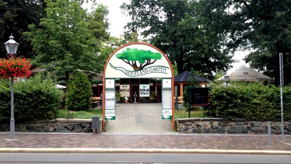 Großer Eingang in den Vogtlandgarten. Links und rechts sind Bäume, darunter Sonnenschirme. Vorne im Bild Fußweg, davor Straße