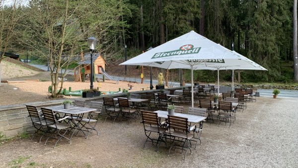 Sitzbänke unter Sonnenschirm. Dahinter Spielplatz und Wald