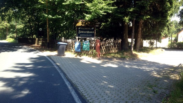 Angebotsschilder und Mülltonne an Zaun unter Bäumen. Davor abzweigende Grundstückseinfahrt nach rechts von Straße.