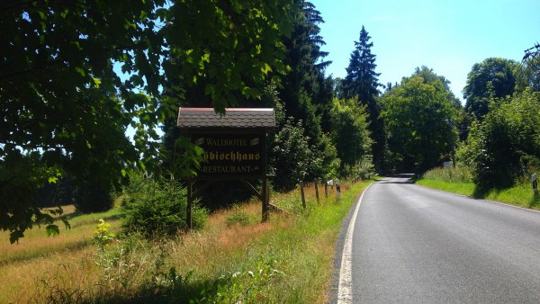 Landstraße neben Wiese bei schönem Wetter. Zwischen Bäumen steht das Willkommensschild zum Zöbischhaus